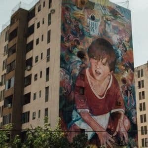 Un enorme murale colorato di un giovane ragazzo con una maglietta rossa è dipinto sul fianco di un condominio a più piani.