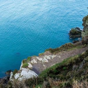 Ein schmaler Feldweg führt entlang einer rauen, grasbewachsenen Klippe mit Blick auf das blaue Meer.