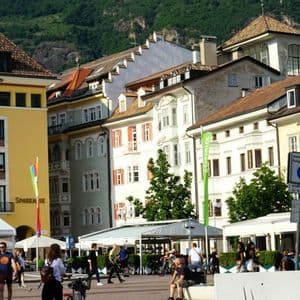 Menschen spazieren durch einen sonnigen europäischen Stadtplatz mit einer markanten weißen Statue, farbenfrohen Gebäuden und einem Berg im Hintergrund.