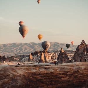 Hot air balloons float over a landscape of unique rock formations as a group of people watches from the ground.