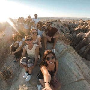Un viaje en grupo de WeRoad se toma una selfie en una cima rocosa, con un vasto cañón de formaciones rocosas al fondo al atardecer.