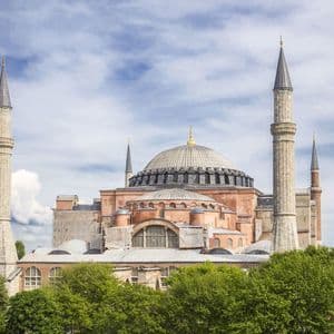 Una grande moschea con una cupola centrale e alti minareti si erge sopra le cime degli alberi lussureggianti sotto un cielo blu con nuvole bianche.