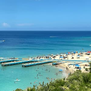 Una vista aérea de una playa concurrida donde la gente nada en aguas turquesas y se relaja en la arena junto a largos muelles azules.