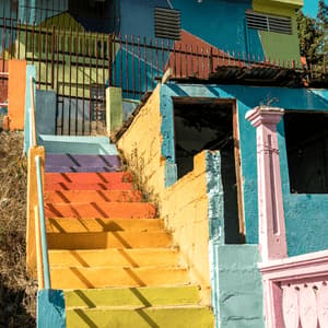 Una escalera exterior pintada con un degradado de arcoíris sube a un edificio colorido bajo un cielo parcialmente nublado.