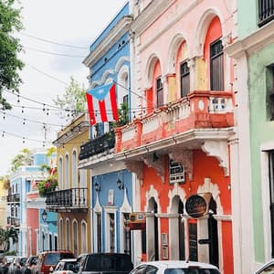 Una bandera puertorriqueña cuelga de un balcón en una calle llena de edificios coloniales coloridos y coches aparcados.