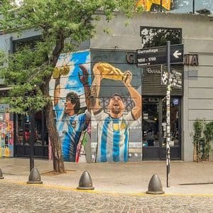 Un grande murale all'angolo di un edificio raffigura i calciatori Maradona e Messi che festeggiano con i trofei della Coppa del Mondo.