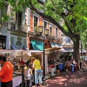 Un vivace mercato all'aperto in una giornata di sole, con persone che curiosano tra le bancarelle sotto alberi frondosi accanto a edifici storici.