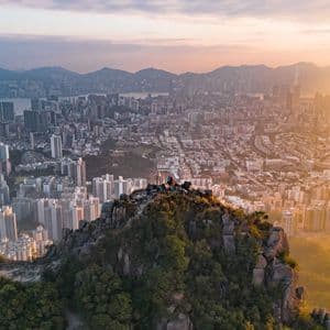 Two people on a rocky mountain peak overlooking a sprawling cityscape at sunrise, with hazy mountains in the background.