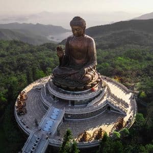An aerial view of a large bronze Buddha statue seated on a tiered platform, surrounded by lush green mountains and distant water.