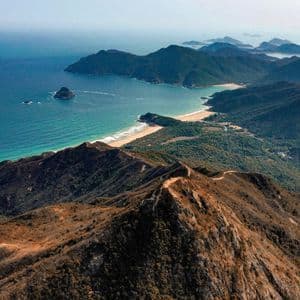 An aerial view from a brown mountain ridge overlooking a coastline with a sandy beach, green hills, and turquoise water under a hazy sky.