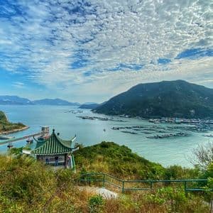 A scenic overlook with a green-roofed pavilion, viewing a bay filled with fish farms and surrounded by lush green mountains under a cloudy sky.