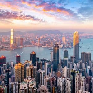 An aerial view of a dense urban skyline with skyscrapers bordering a body of water under a colorful, cloudy sky at sunset.