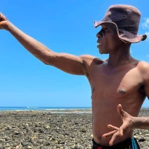 Un uomo con un cappello a secchiello mostra un polpo appena pescato su una spiaggia rocciosa sotto un cielo azzurro e limpido.