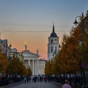 Une large rue bordée d'arbres avec un feuillage d'automne mène à une cathédrale et un clocher au crépuscule, avec des gens marchant au loin.