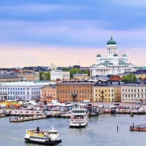 An aerial view of a bustling city harbor with ferries, waterfront buildings, and a prominent white cathedral with green domes in the background.