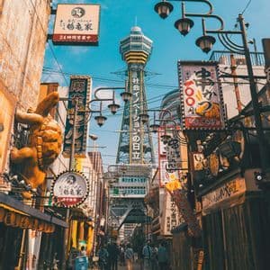 A view down a narrow city street crowded with Japanese signs, with a tall observation tower in the distance under a blue sky.
