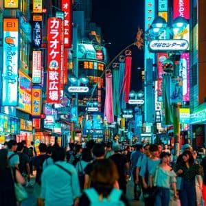 A large crowd of people walks down a narrow city street at night, illuminated by many colorful neon signs.