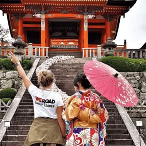 A WeRoad group leader makes a peace sign next to a woman in a kimono at the base of stairs leading to a Japanese temple.