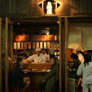 Two women on a WeRoad group trip dining at a traditional Japanese restaurant at night, viewed from the outside.