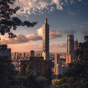 Un alto grattacielo a gradoni si staglia sullo skyline di una città al tramonto, visto da una foresta scura e rigogliosa.