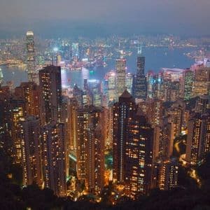 Una veduta aerea di un paesaggio urbano denso con grattacieli illuminati che circondano un porto al tramonto, con montagne sullo sfondo.