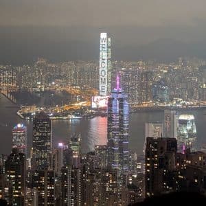 Uno skyline cittadino esteso visto da un punto di osservazione elevato di notte, con un grattacielo che mostra la parola 'BENVENUTI' in luci verticali.