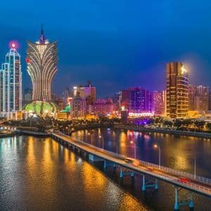 Uno skyline cittadino moderno con grattacieli illuminati di notte, con un ponte che attraversa uno specchio d'acqua in primo piano.