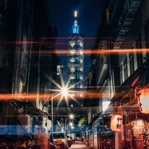 Un grattacielo alto e illuminato visto da uno stretto vicolo cittadino di notte, con scooter parcheggiati, lanterne luminose e scie luminose.