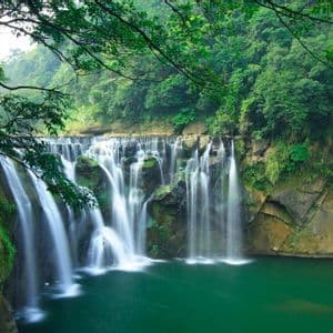 Una vasta cascata con flussi d'acqua a lunga esposizione si riversa in una piscina turchese, circondata da una foresta lussureggiante e verde.