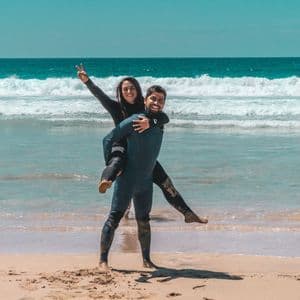 Un uomo in muta porta in spalla una donna sorridente su una spiaggia sabbiosa, con onde turchesi dell'oceano dietro di loro.