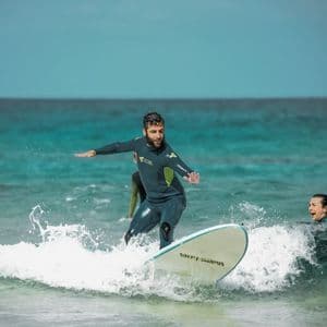 Un uomo del gruppo WeRoad, in muta, sta su una tavola da surf cavalcando un'onda, mentre altri osservano dall'oceano turchese.