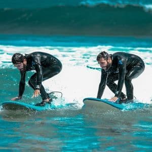 Due uomini in mute nere di un viaggio di gruppo WeRoad surfano affiancati su tavole blu nell'oceano.