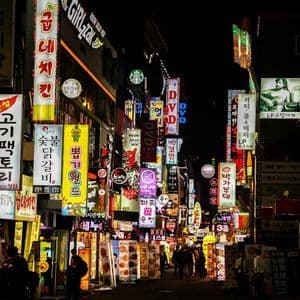 Una strada cittadina affollata di notte, illuminata da molte insegne al neon verticali e colorate, con scritte in coreano.