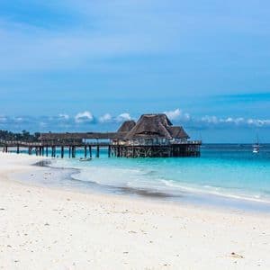 Ein Holzgebäude mit Reetdach auf Stelzen steht am Ende eines Stegs über türkisfarbenem Wasser, neben einem weißen Sandstrand.