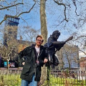 Un uomo sorridente con gli occhiali posa accanto a una statua di bronzo di una figura che vola su una scopa in una piazza della città.