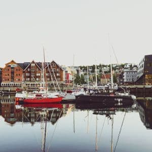 Boote liegen in einem ruhigen Hafen vor Anker, während sich traditionelle und moderne Gebäude entlang der Uferpromenade im Wasser spiegeln.