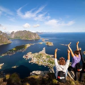 A WeRoad group trip of three women sitting on a clifftop, arms raised, overlooking a coastal village surrounded by mountains and sea.