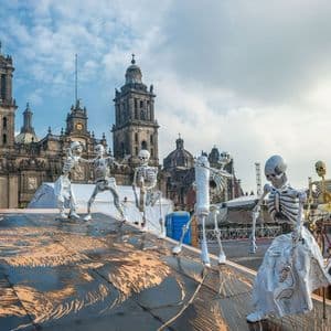 Varias figuras de esqueletos grandes para un festival se encuentran en una plaza frente a una gran catedral de dos torres bajo un cielo azul.