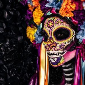 Una persona di profilo con trucco da teschio di zucchero paillettato e un grande copricapo floreale colorato su sfondo scuro.