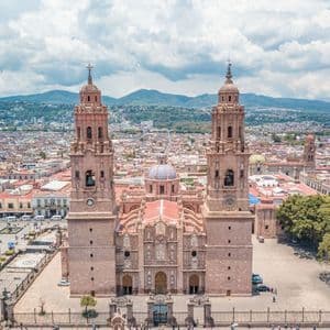 Una vista aerea di una grande e ornata cattedrale in pietra con due campanili e una cupola centrale, situata in una piazza cittadina con montagne in lontananza.
