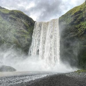 Una potente cascada cae por un acantilado verde y cubierto de musgo sobre una oscura orilla de guijarros, creando una densa niebla bajo un cielo nublado.