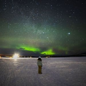 Une personne vue de dos se tient dans un paysage enneigé la nuit, regardant les aurores boréales vertes et un ciel étoilé.