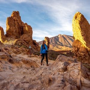 Eine Frau in einer blauen Jacke wandert auf einem felsigen Pfad zwischen großen, sonnenbeschienenen Felsformationen mit einem Berg im Hintergrund.
