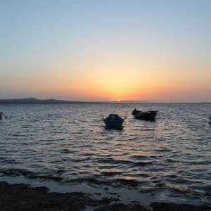 Diverse piccole barche sono ancorate su acque calme al tramonto, con il sole che risplende all'orizzonte dietro una terra lontana.