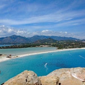 Una vista panoramica di un paesaggio costiero con kitesurfer su acqua turchese, accanto a una spiaggia di sabbia bianca e una catena montuosa in lontananza.