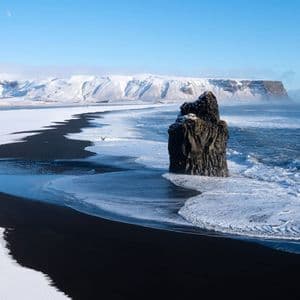 Les vagues déferlent sur une plage de sable noir, partiellement recouverte de neige, avec un imposant piton rocheux au large et des montagnes enneigées en arrière-plan.