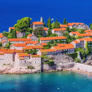 Veduta dall'alto di un villaggio isolano con case in pietra e tetti rossi, delimitato da una spiaggia sabbiosa e dal mare blu.