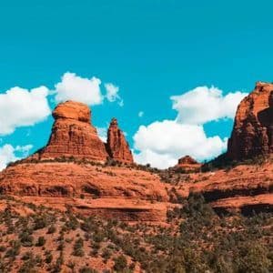 Geschichtete rote Felsformationen erheben sich aus einer Landschaft grüner Sträucher unter einem strahlend blauen Himmel mit bauschigen weißen Wolken.
