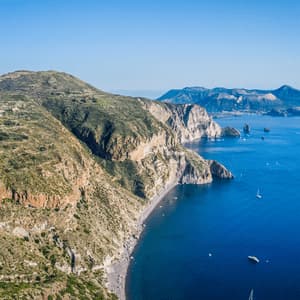 Vue aérienne d'un littoral accidenté et verdoyant rencontrant une mer d'un bleu profond, avec une plage de galets sombres et plusieurs voiliers dans la baie.