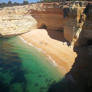 Ein abgelegener Sandstrand mit klarem, türkisfarbenem Wasser, umgeben von hohen Klippen mit Höhlen, aus der Vogelperspektive gesehen.